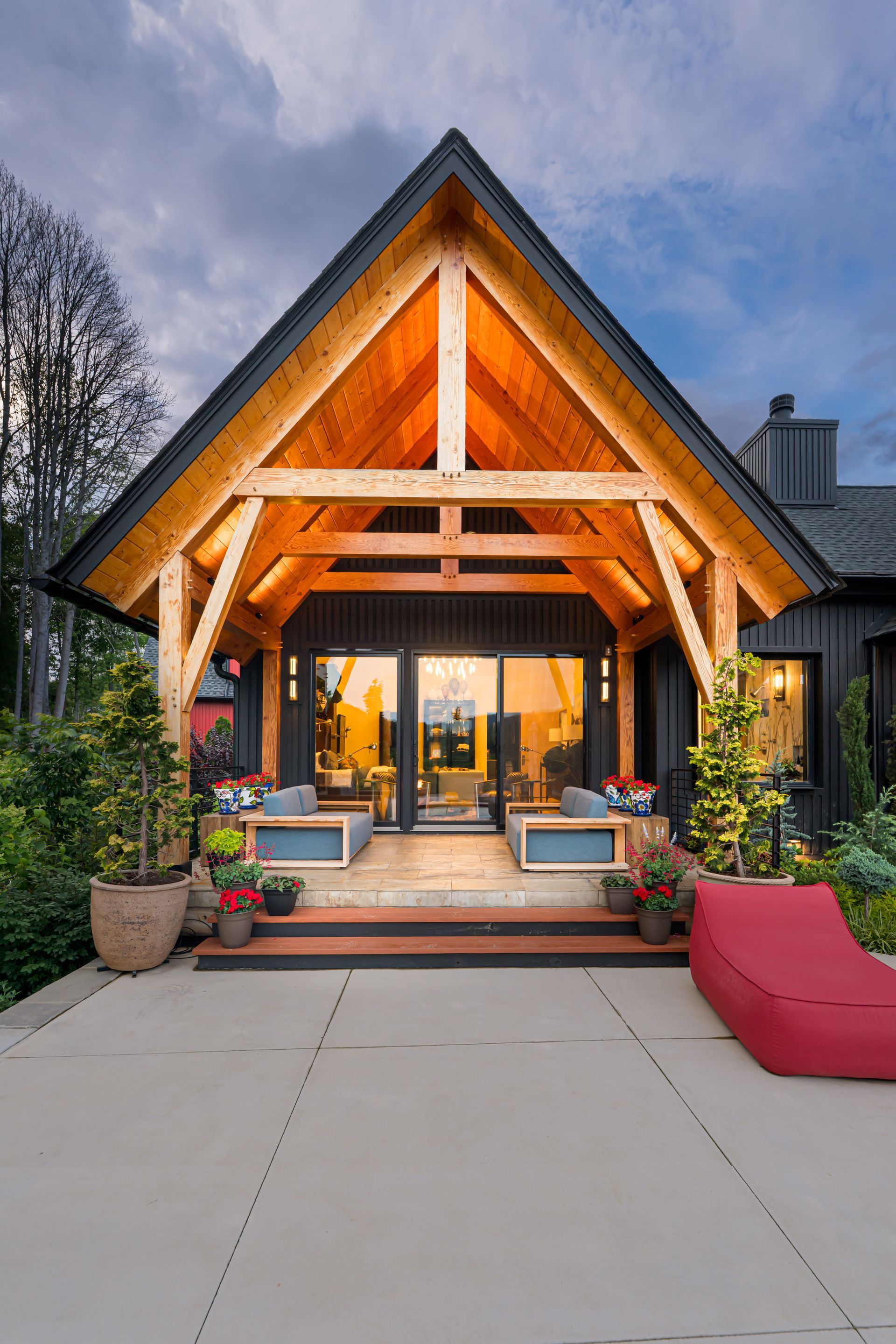 Modern chalet-style house entrance with warm-lit gable porch, patio seating, and potted plants at dusk