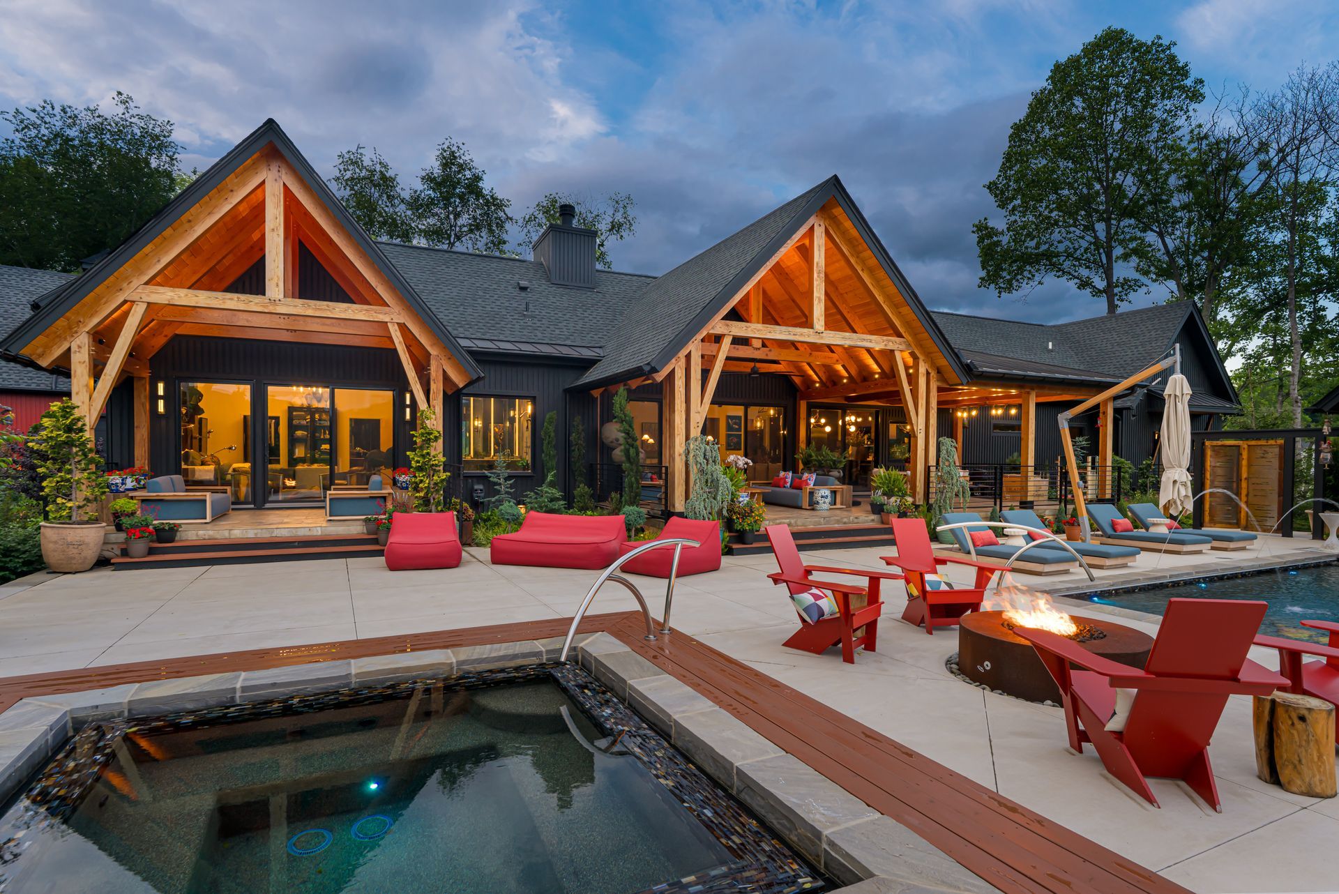 Modern lodge with peaked roofs, warm lights, patio seating, and a pool at dusk