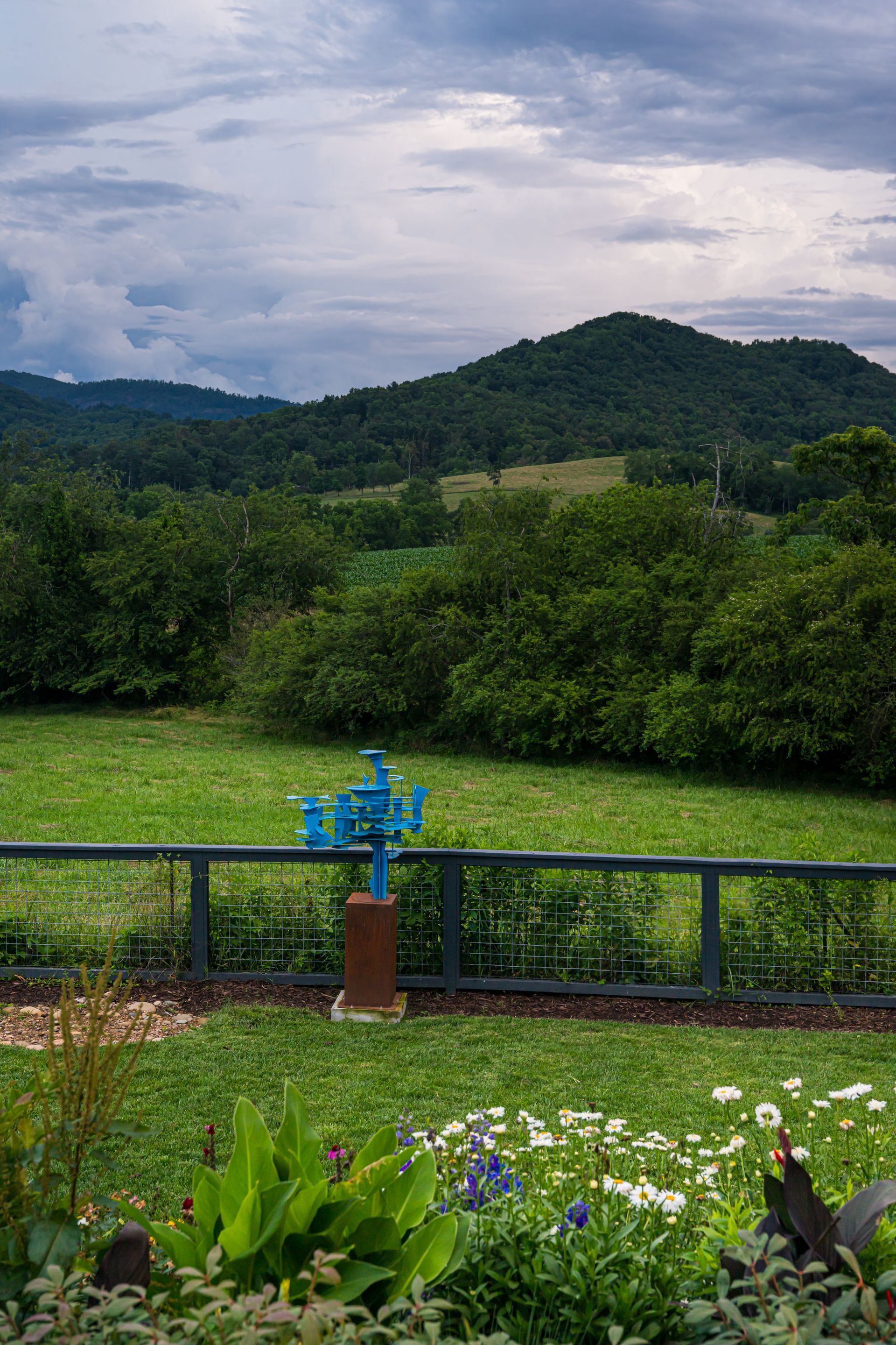 Lush green garden overlooking forested hills under a cloudy sky, with a blue sign in the foreground