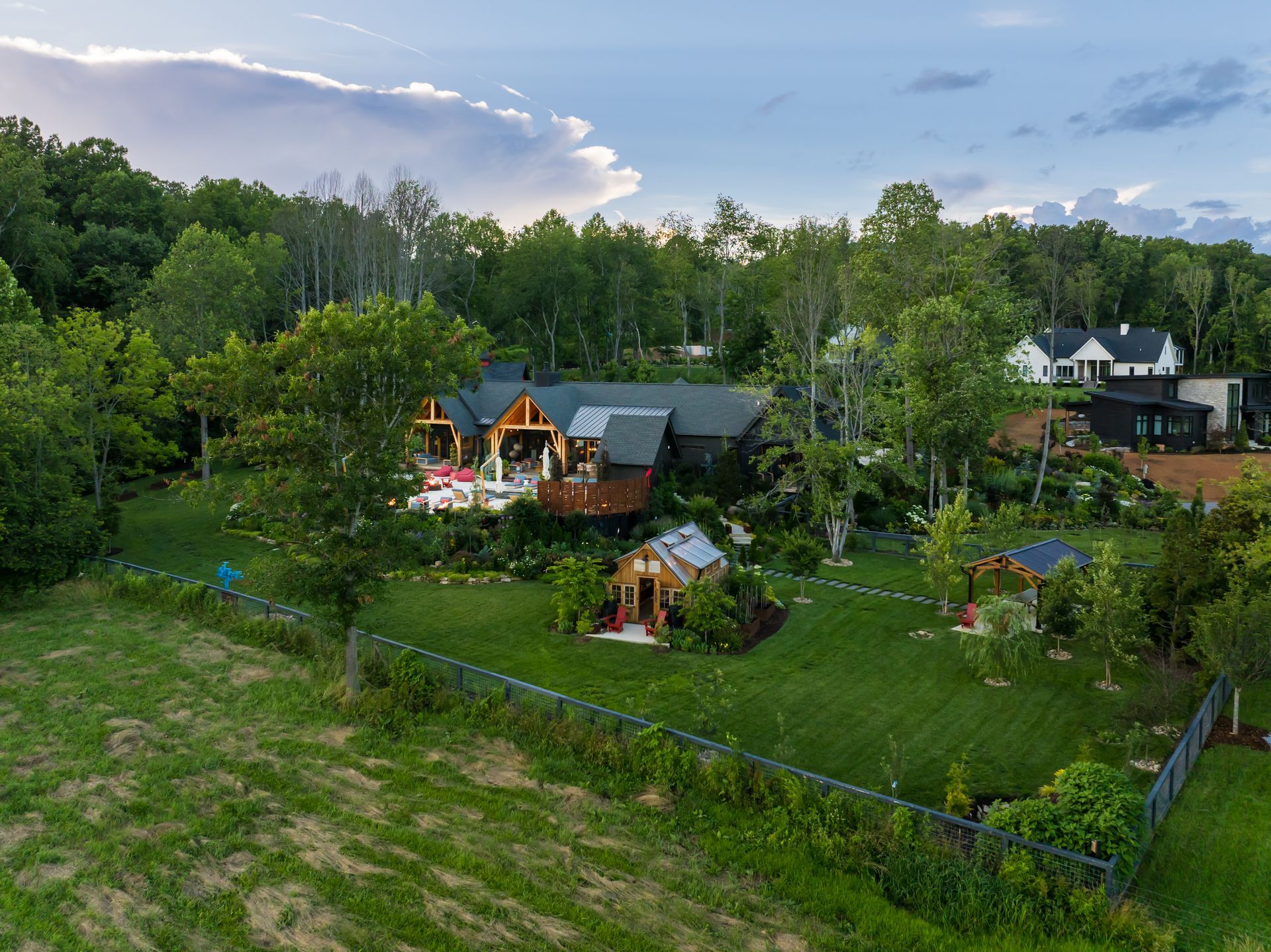 Rural garden with two small wooden houses, lush green trees, and a cloudy evening sky
