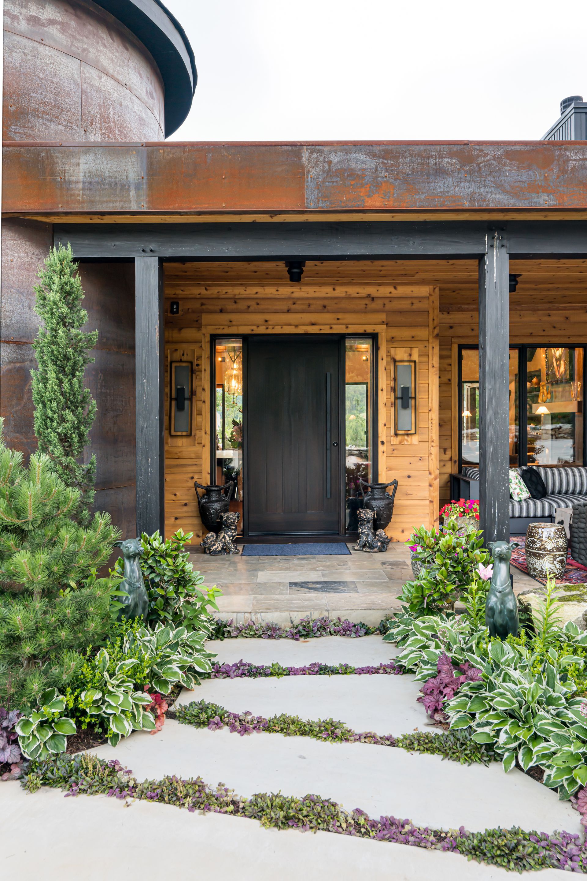 Front porch with dark door, warm wood siding, and stone path bordered by lush green plants