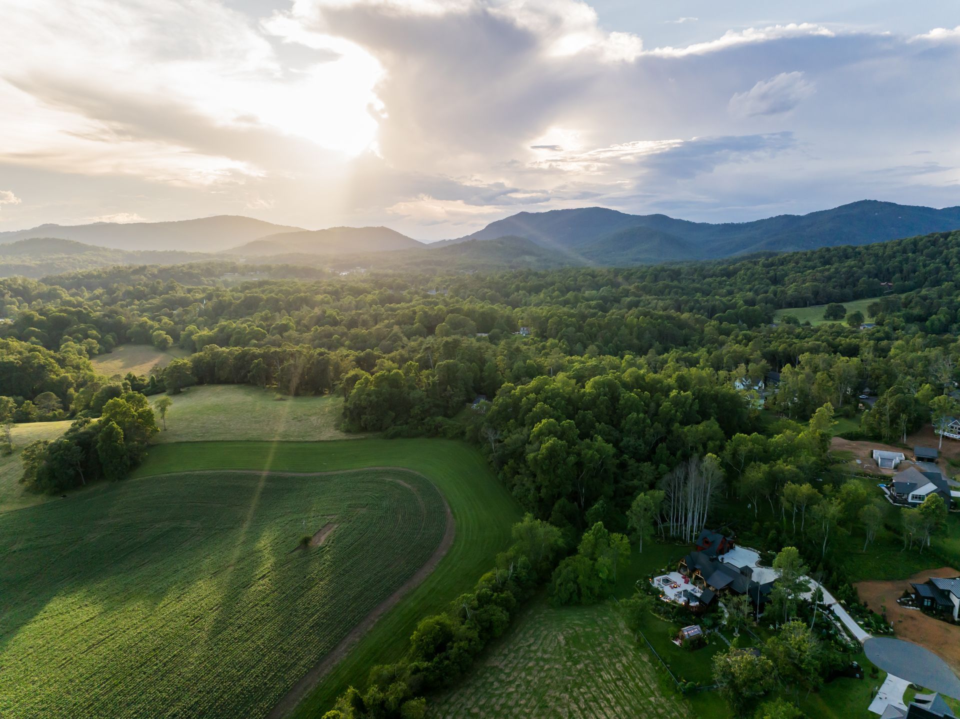 Sunlit rural valley with green fields, forested hills, and scattered houses under a cloudy sky