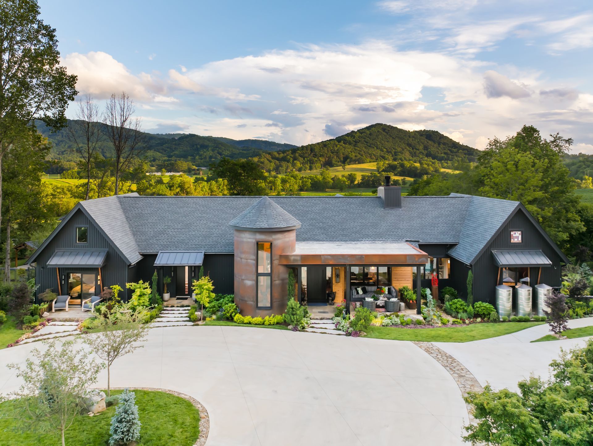 Modern single-story house with gray roof, stone facade, and landscaped driveway in a green mountain setting