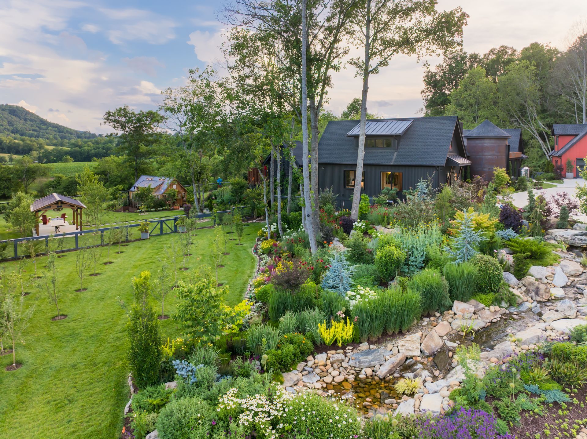 Lush hillside garden with flowers, trees, and cabins at sunset