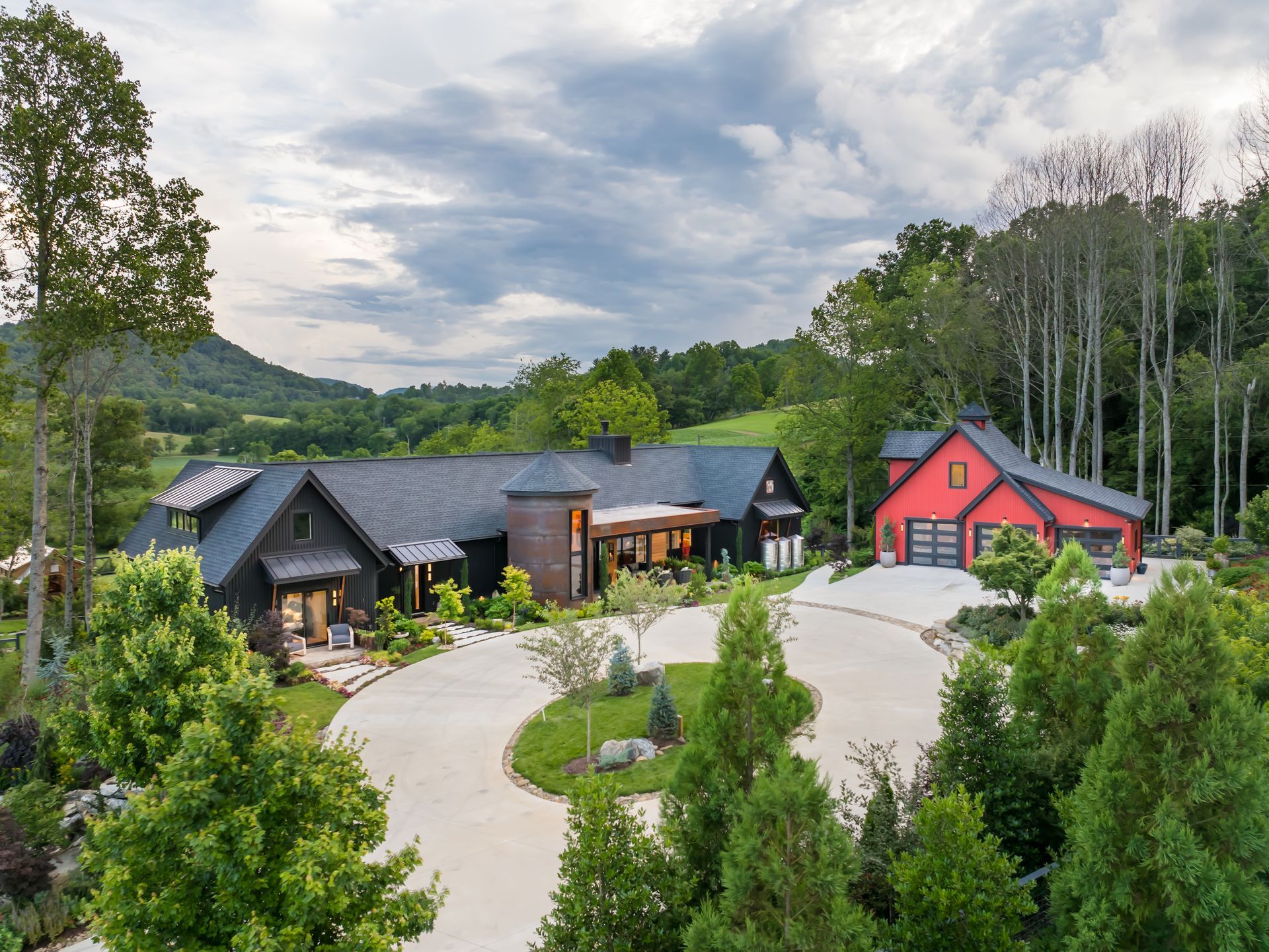 Rustic lodge buildings with gray and red roofs beside a curving driveway, surrounded by trees and hills