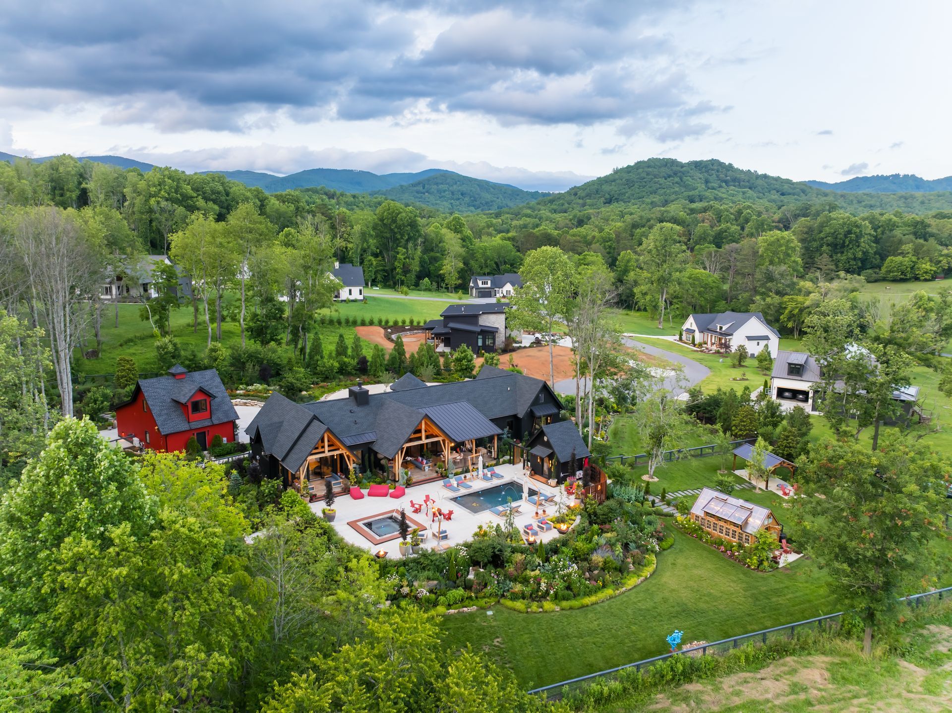 Aerial view of a rustic resort with black-roof cabins, pool, and tennis court amid lush green hills.