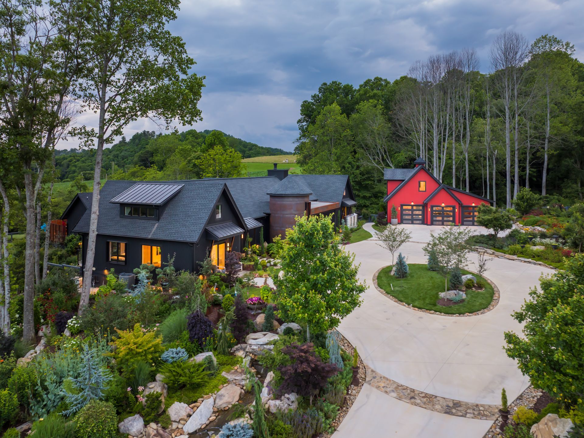 Modern house and red barn-style building in a landscaped rural courtyard with stone paths and trees