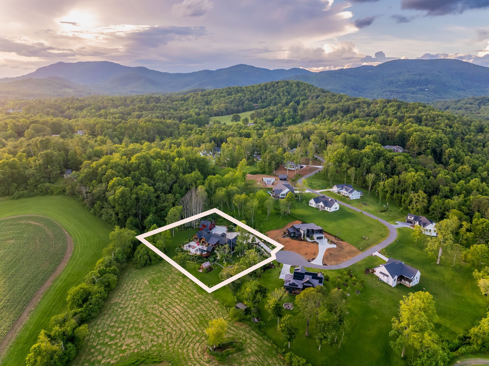 Aerial view of a rural neighborhood with houses, green fields, and wooded hills at sunset.