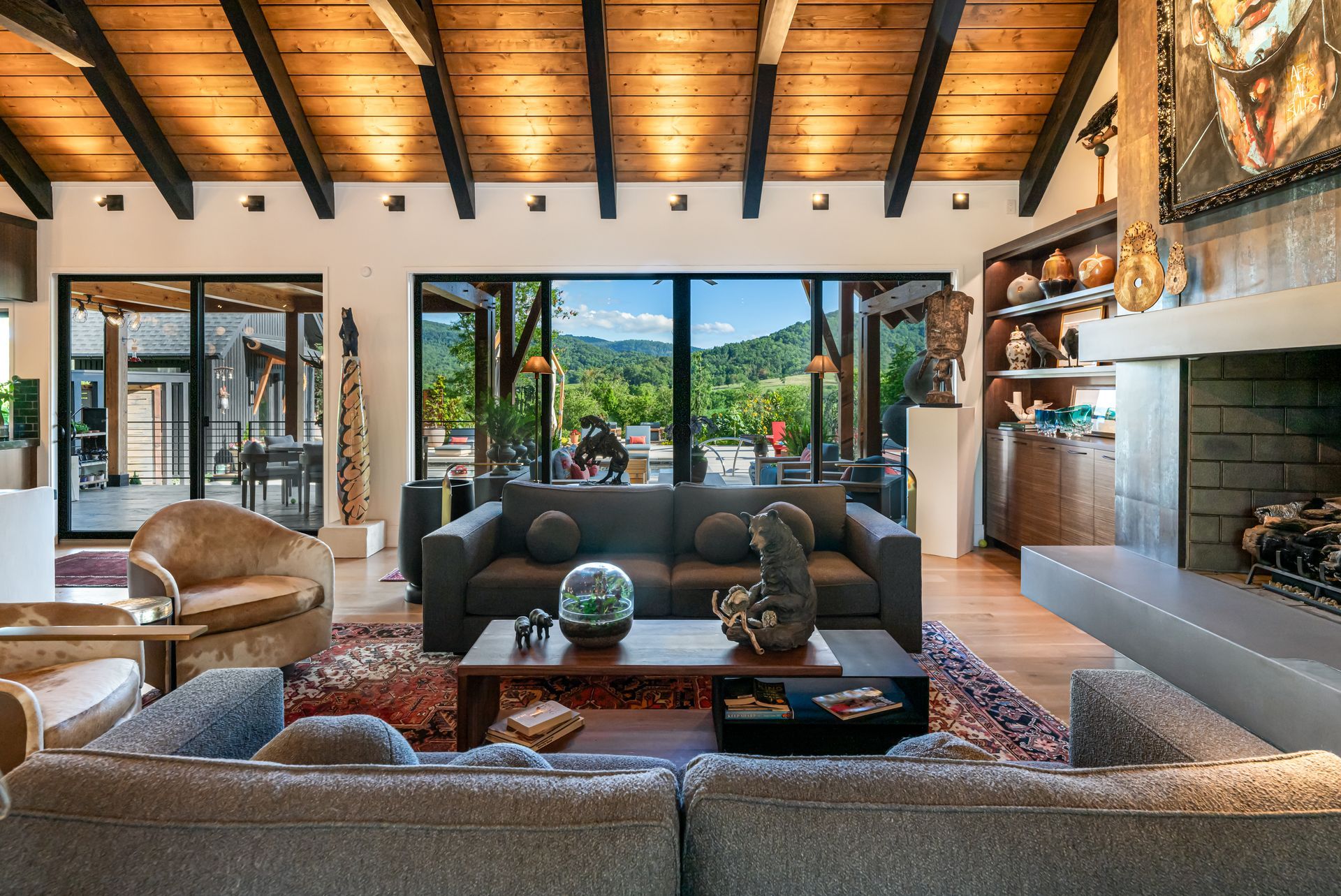 Modern living room with wood beam ceiling, stone fireplace, sofas, and large open doors to a mountain view