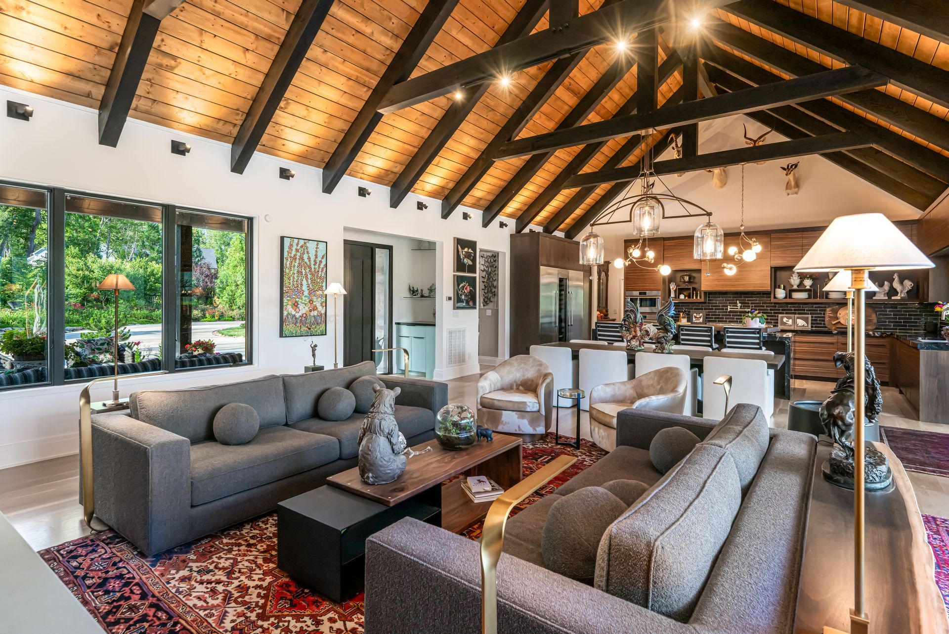 Open-plan living room with gray sofas, vaulted wood ceiling, large windows, and kitchen in the background