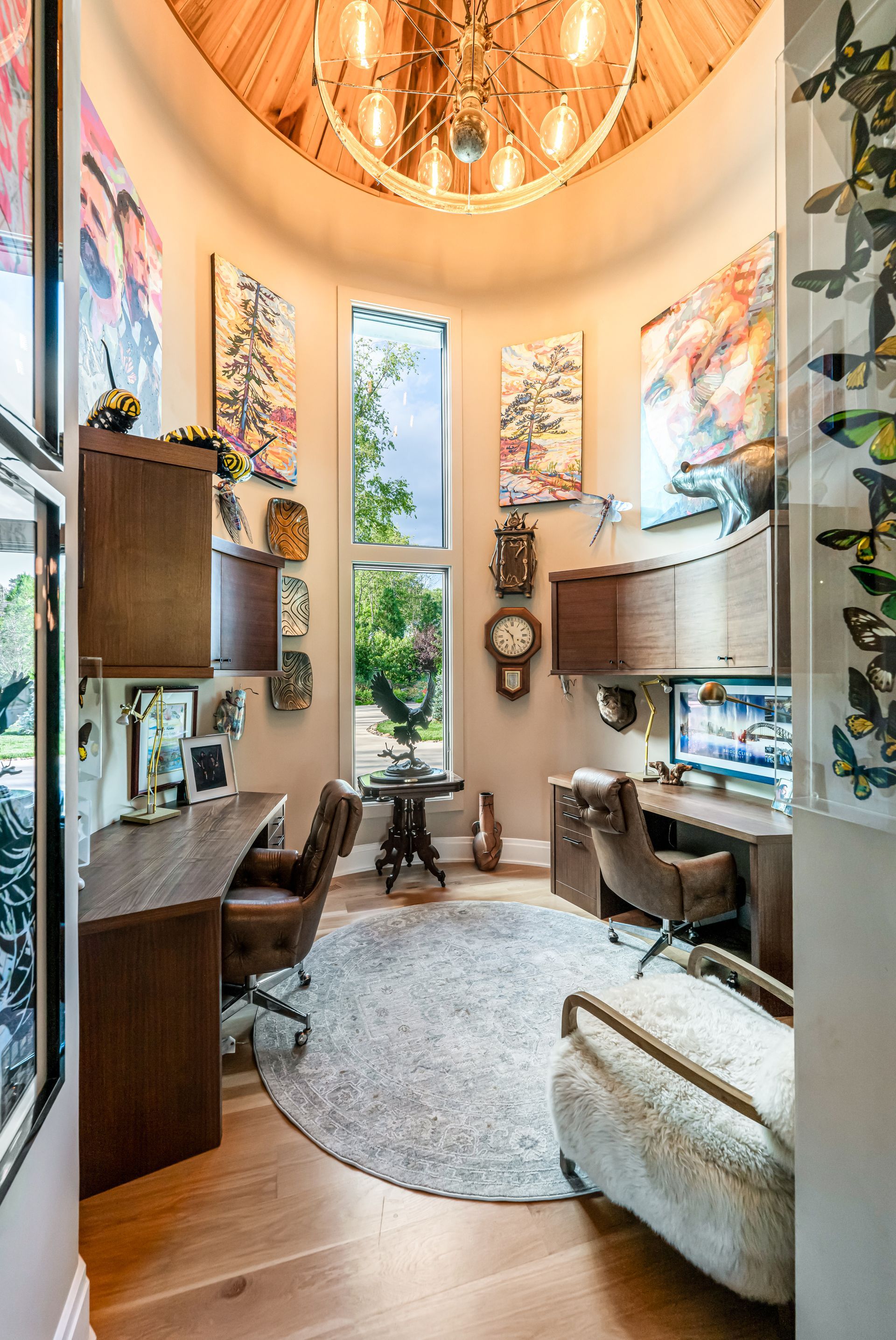 Tall, bright living room with high windows, plants, shelves, a desk, and a cozy chair under warm wood ceiling lines
