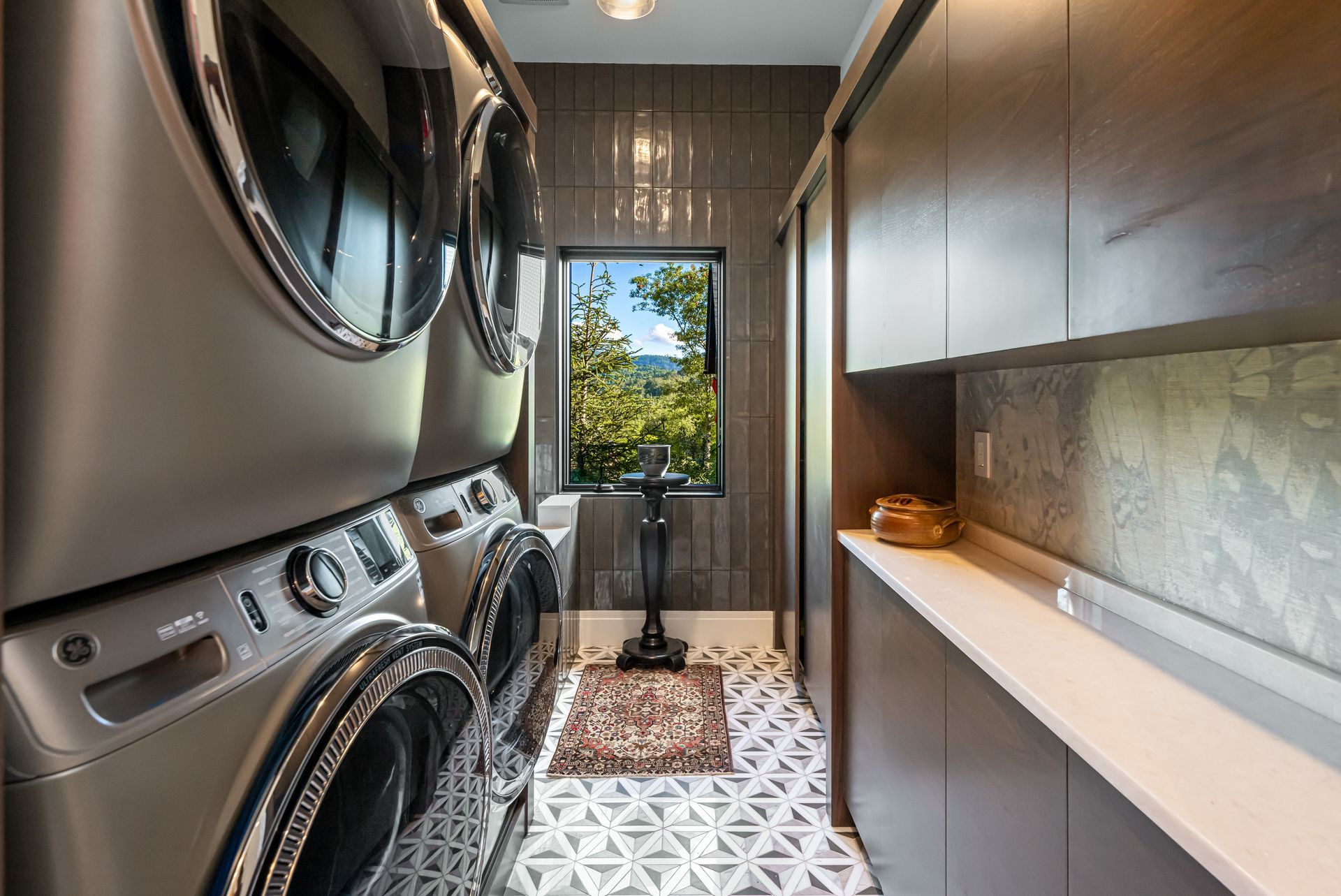 Narrow laundry room with stacked washer and dryer, patterned floor tiles, and a window at the end.