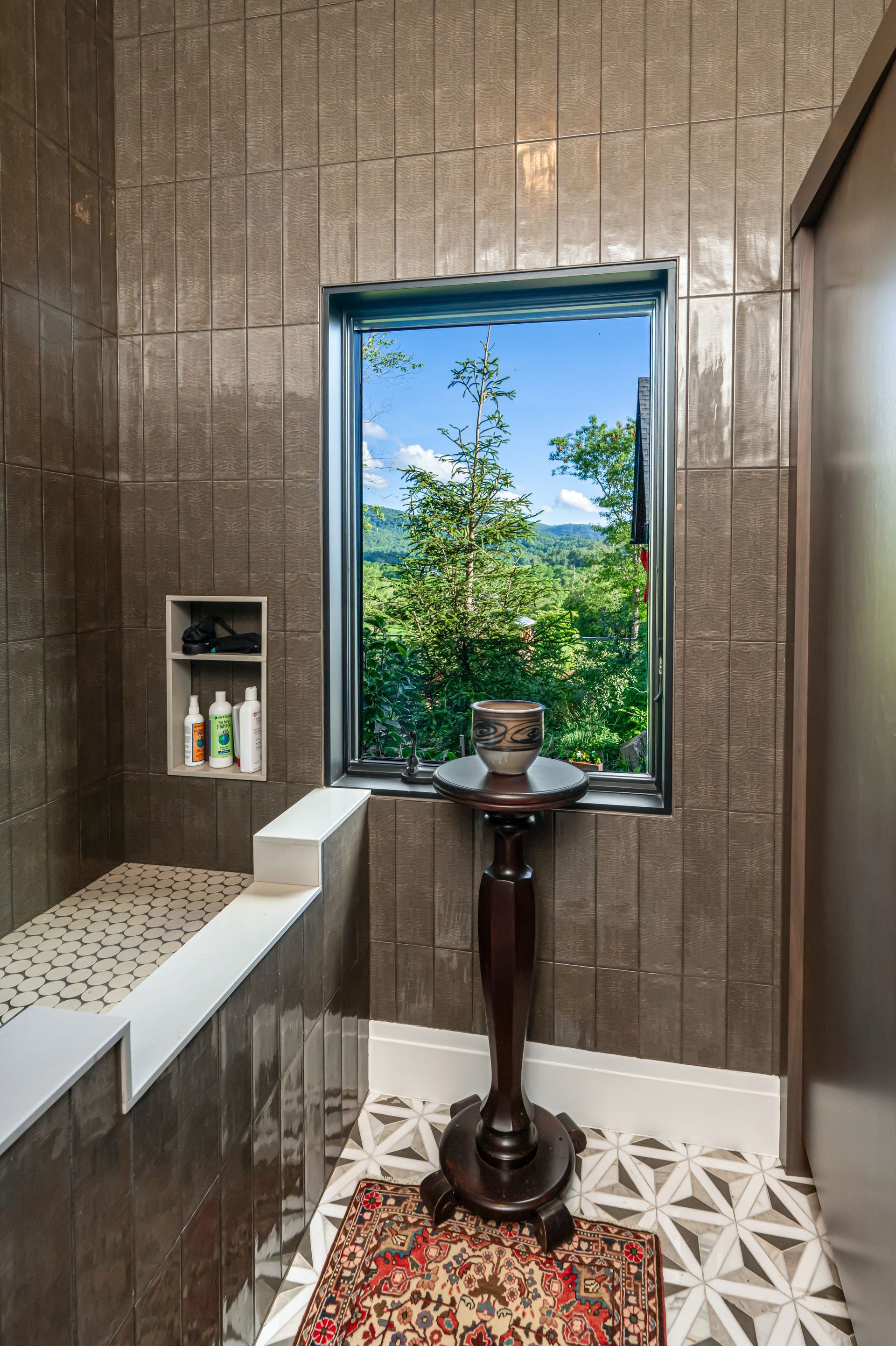 Small tiled bathroom with window, wall shelf, and pedestal sink topped with a basin and flowers.