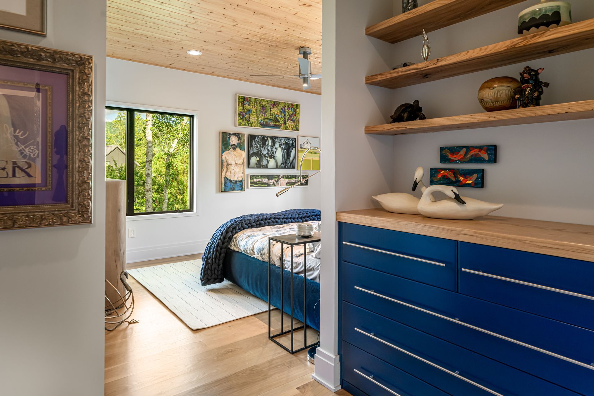 Bright bedroom with blue dresser, bed, shelves, and window overlooking greenery