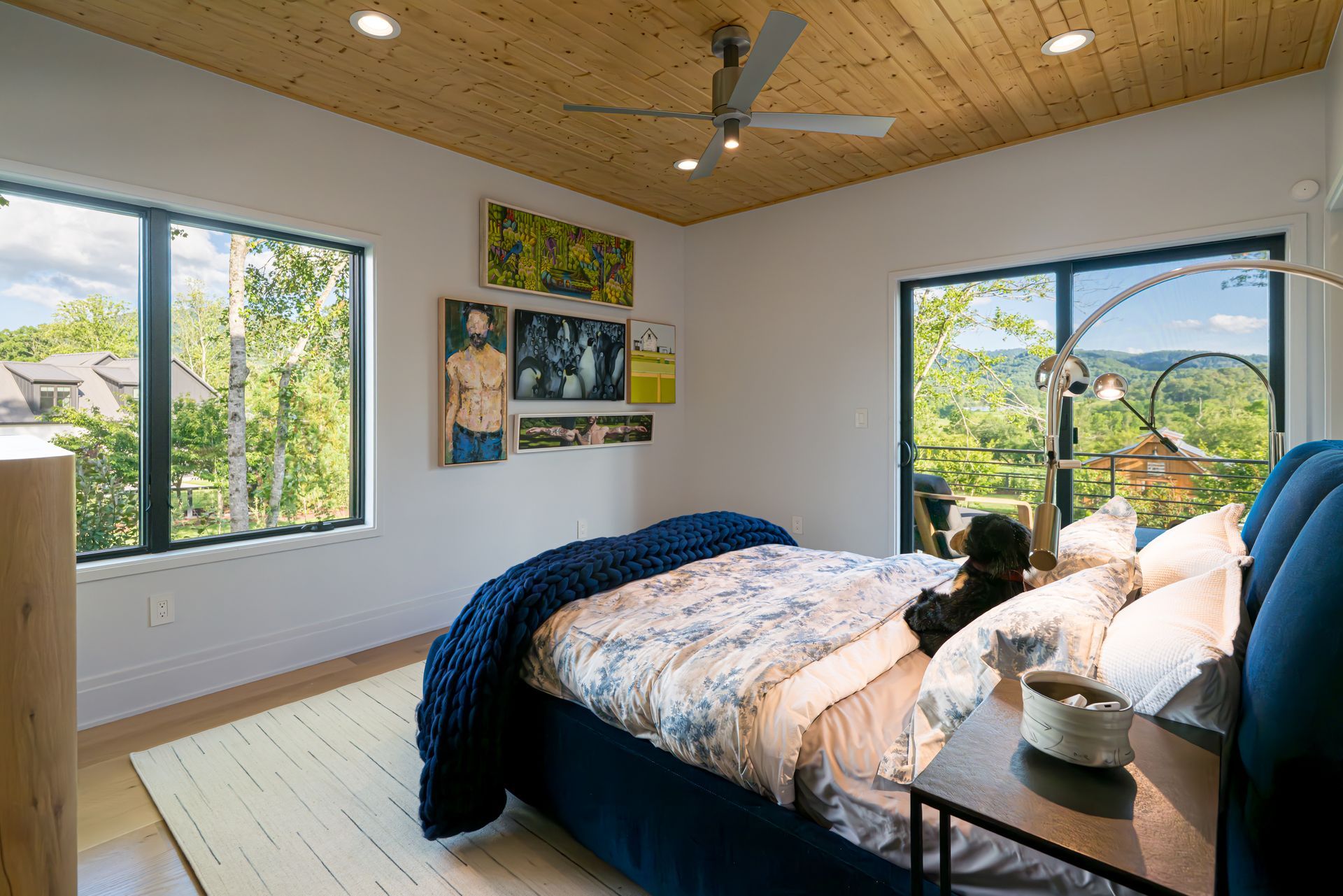 Bright bedroom with a bed, large windows, wood ceiling, and blue accents overlooking greenery