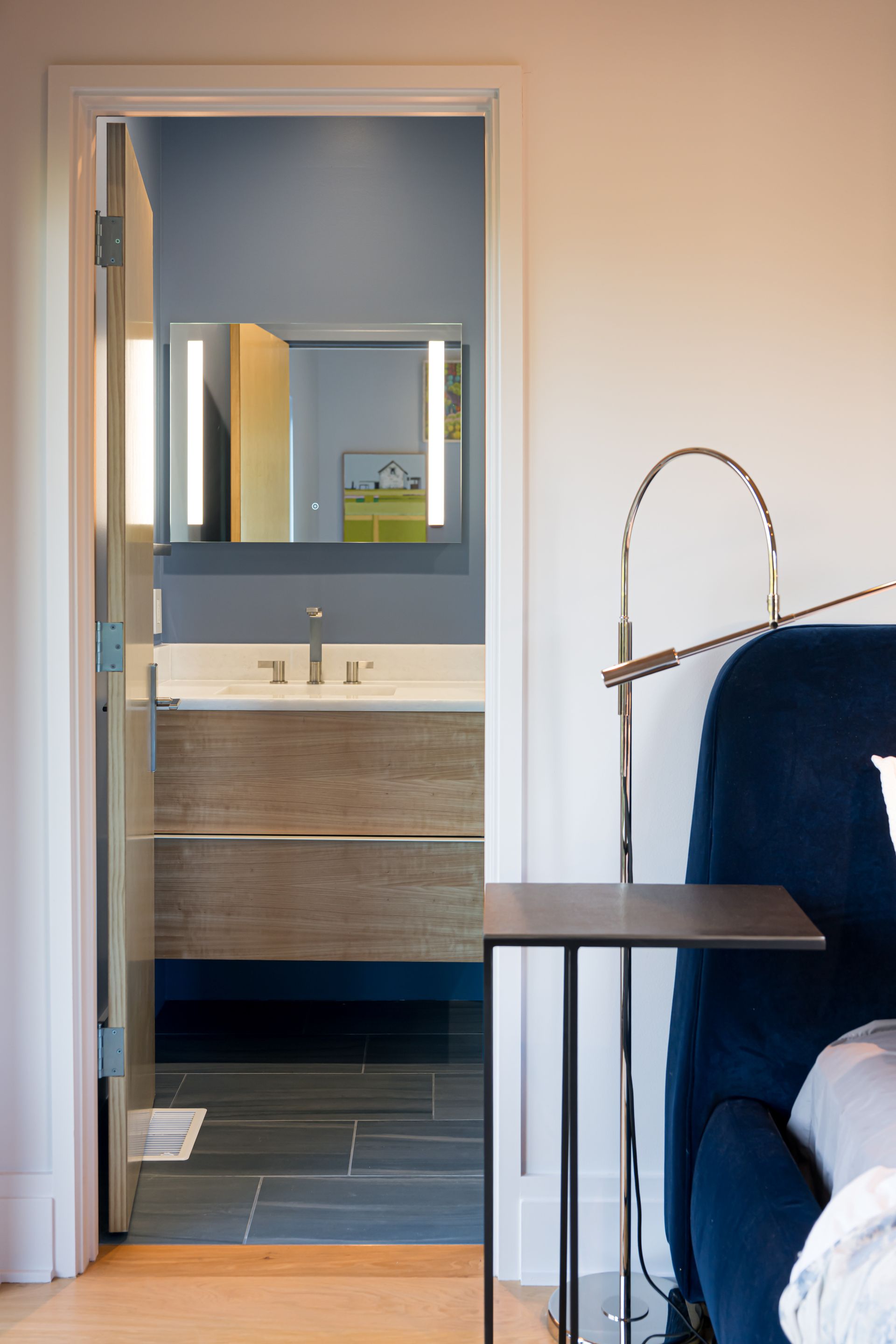 Bedroom view into a bathroom with mirror, sink, wood vanity, and open door beside a blue chair and lamp.