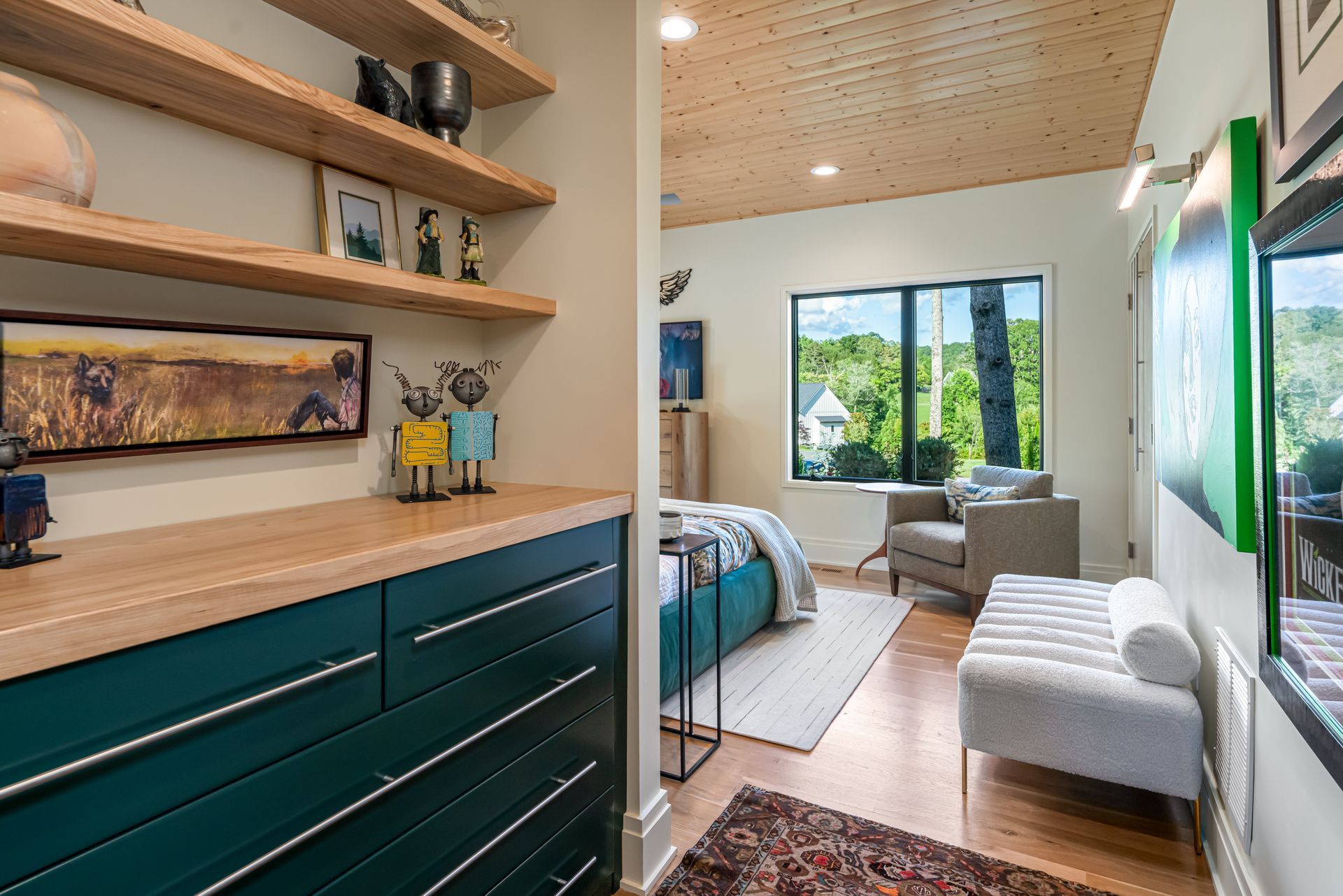 Modern bedroom with teal dresser, shelves, bed, lounge chair, and large window overlooking greenery