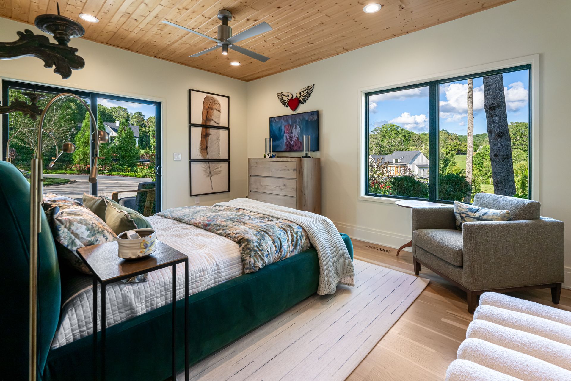 Bright bedroom with large windows, teal bedspread, armchair, and ceiling fan in a tropical view.