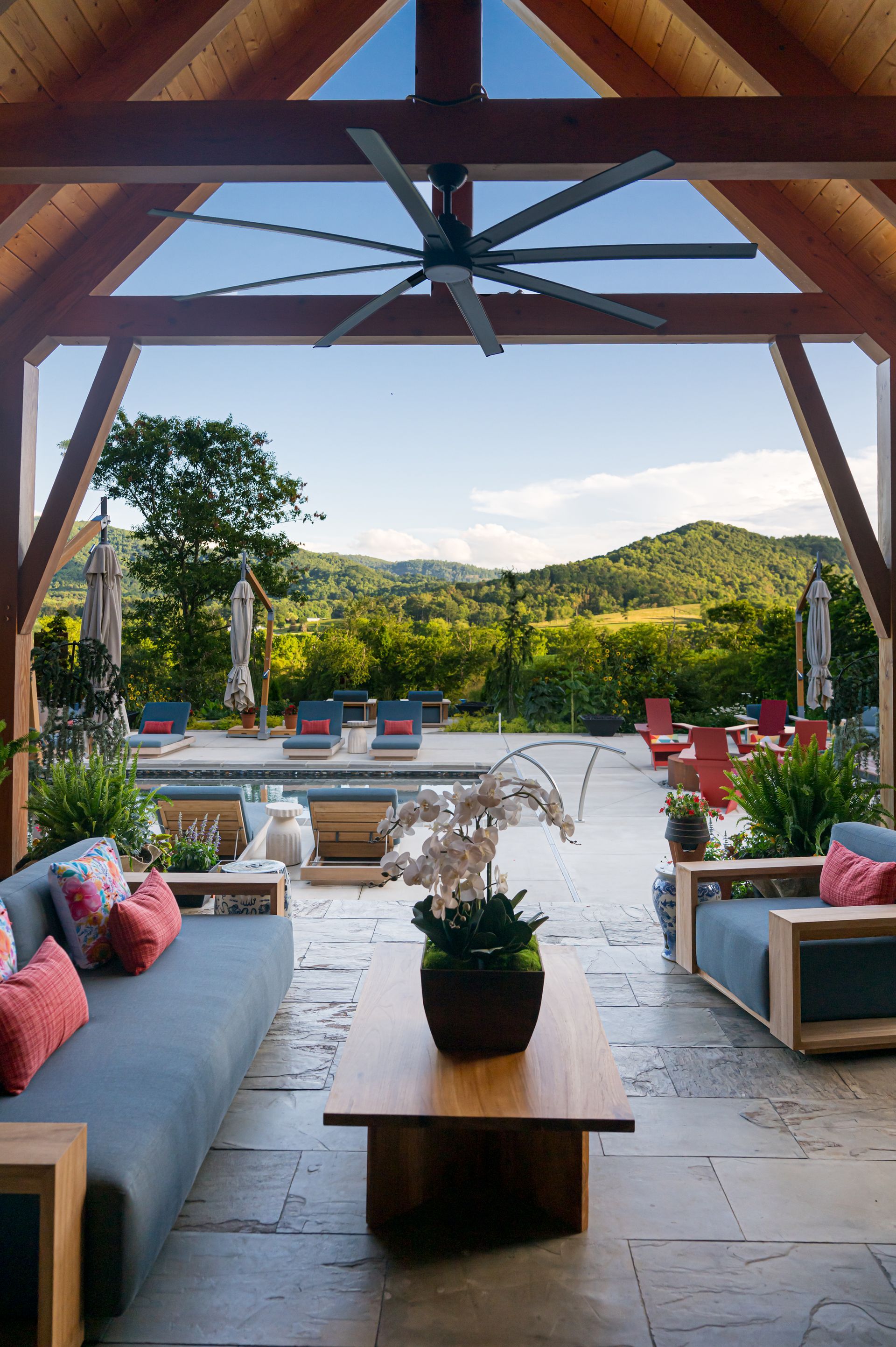 Covered patio lounge overlooking pool and green hills, with gray sofas, wood table, and ceiling fan.