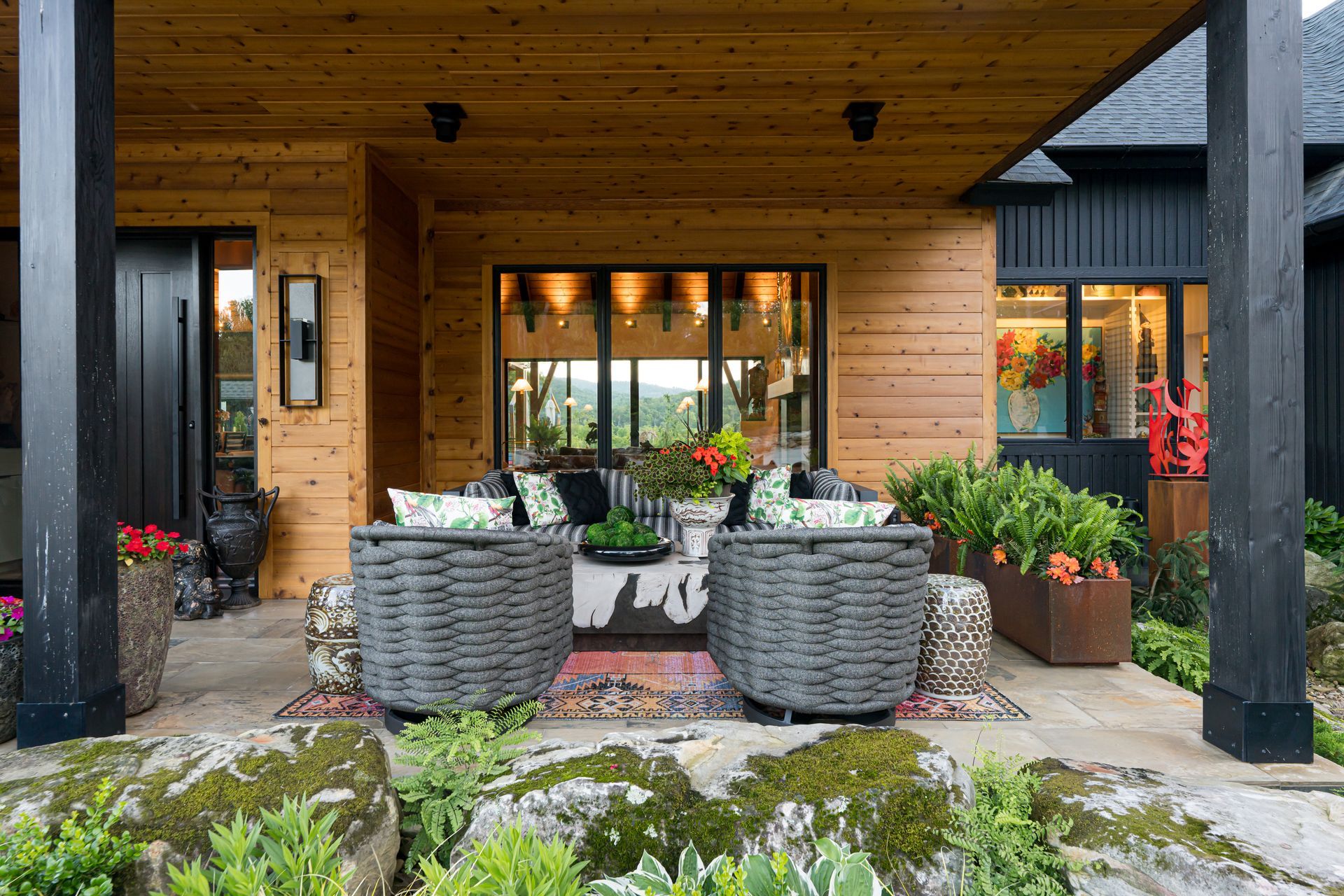 Covered wooden patio with wicker chairs, potted flowers, and glass doors overlooking a garden.