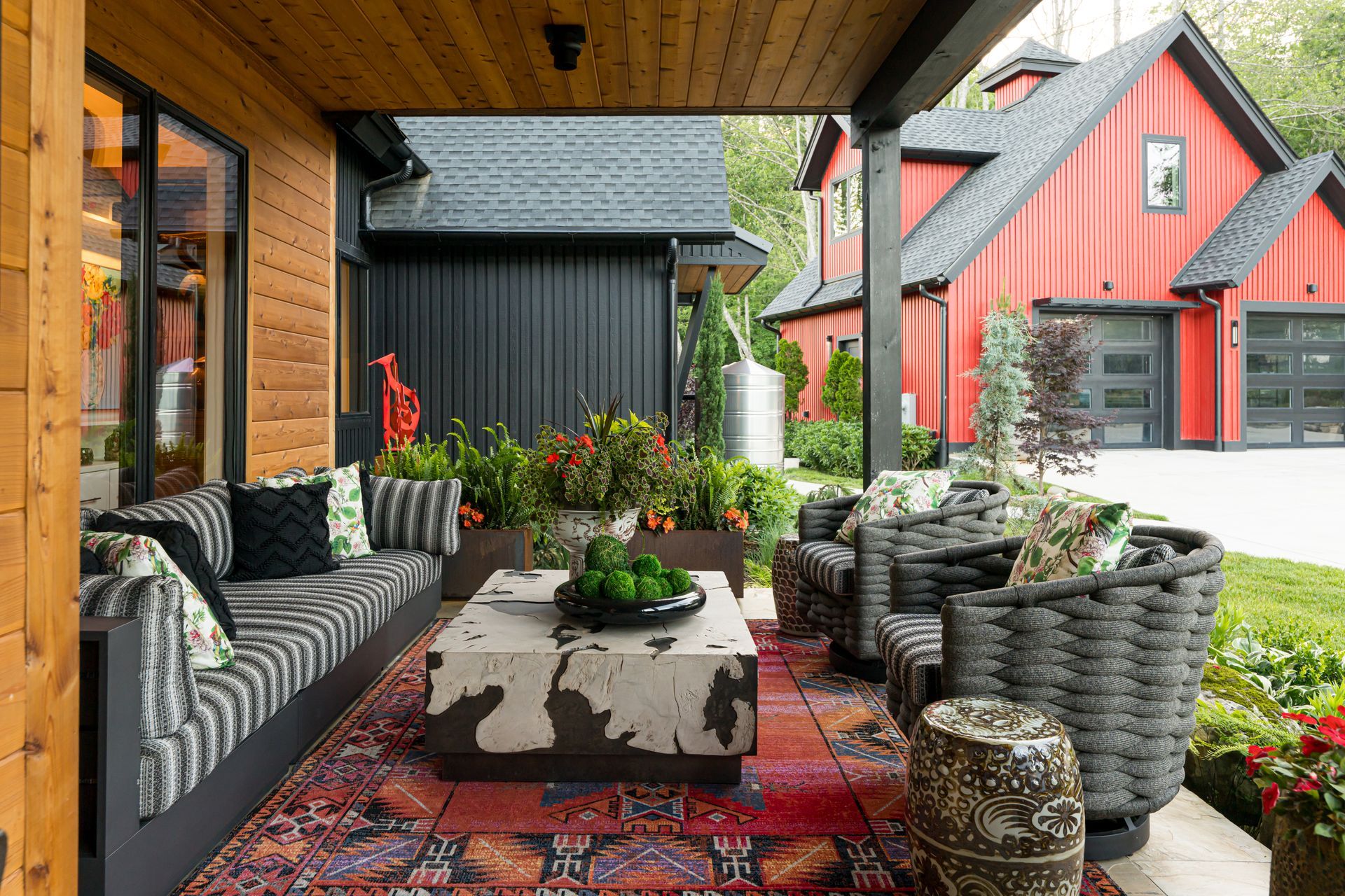 Cozy covered patio with wicker seating, potted plants, and patterned rug beside red house exterior