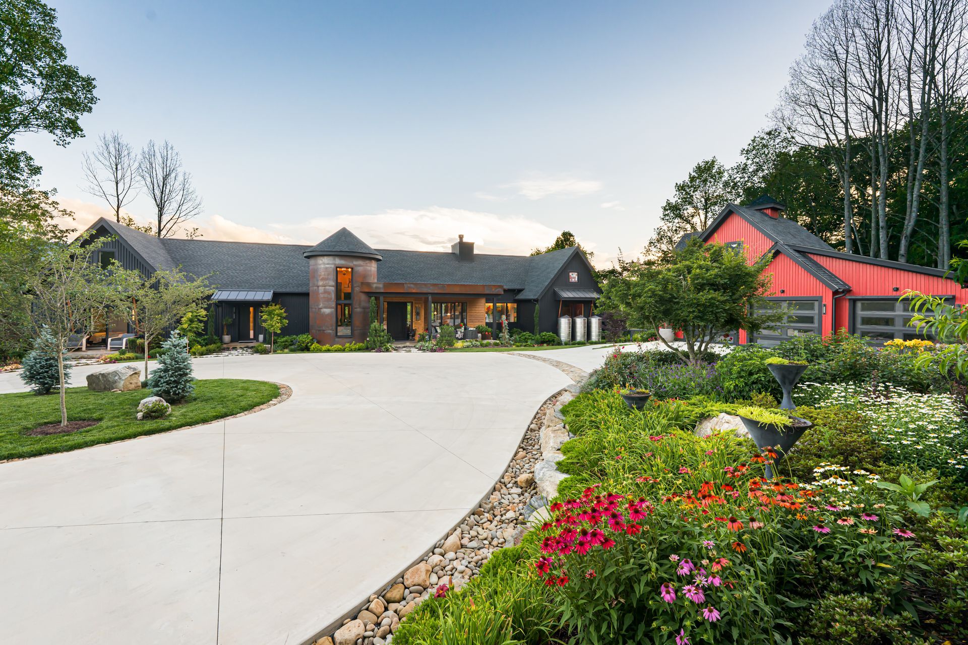Landscaped estate driveway leading to a modern house with flower beds and a red-roofed building nearby