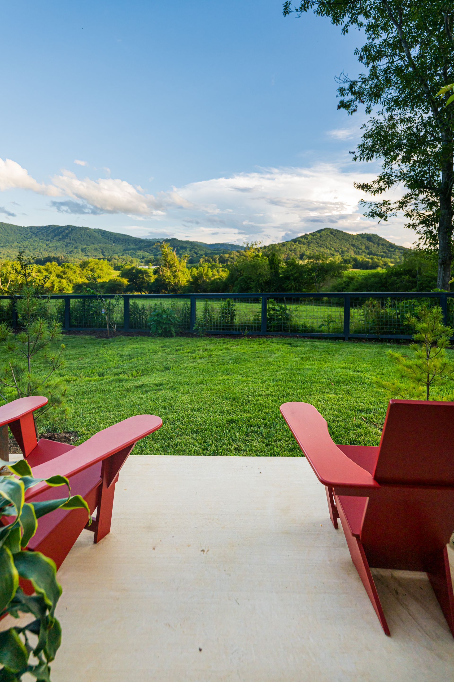 Red chairs on a patio overlooking a green lawn, fence, and sunlit mountains under a blue sky