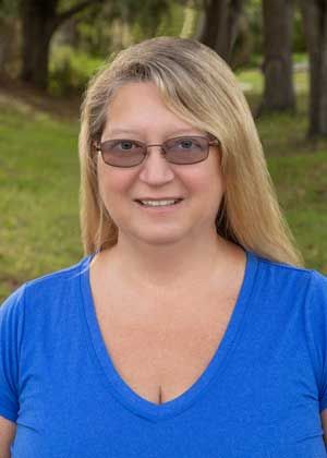 Woman with blonde hair wearing glasses and a blue shirt, smiling outdoors.
