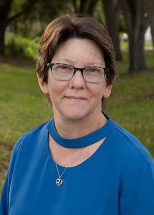 Woman wearing glasses, blue top, and a heart pendant, smiling in an outdoor setting.