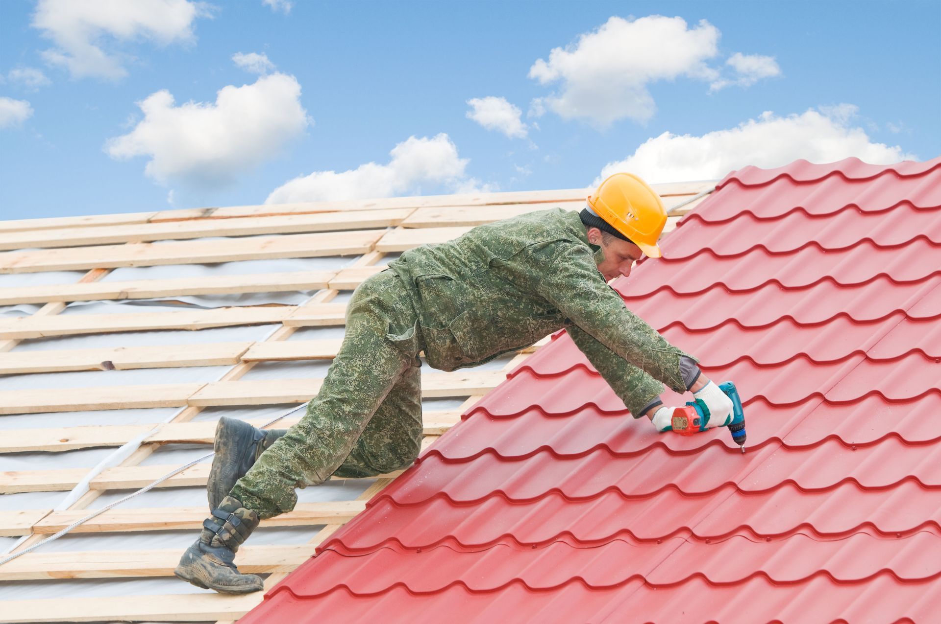 Roofer in camouflage uniform installing red metal roofing on a house under a blue sky with clouds.