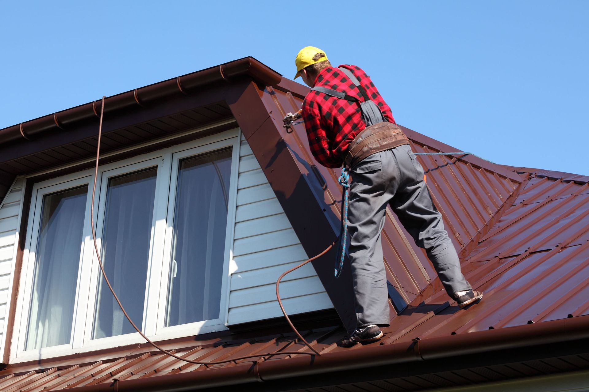 Person in red plaid shirt and safety gear on a brown metal roof, near a white-framed window.