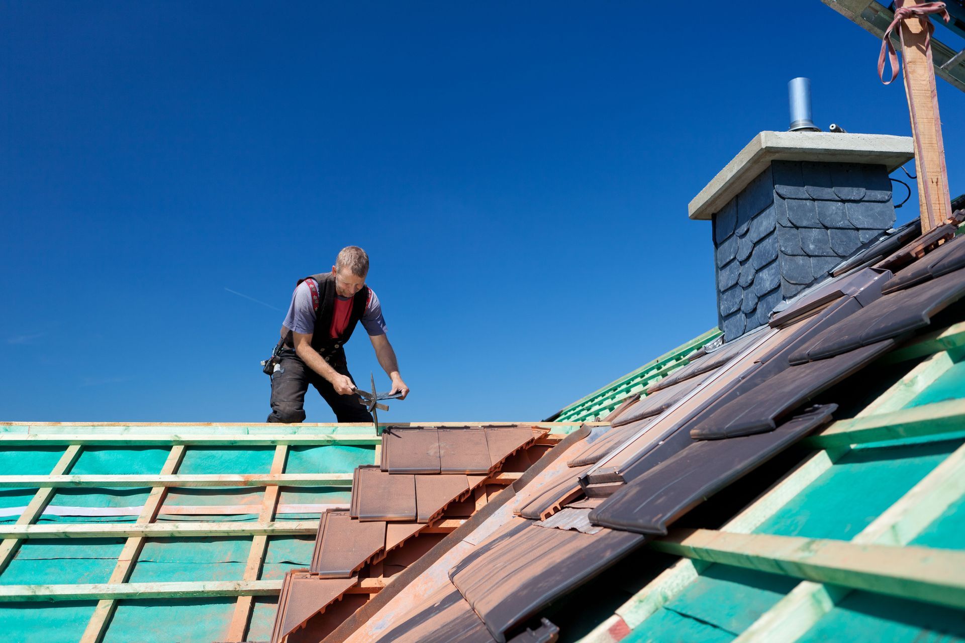 Roofer installing tiles on a roof. Blue sky, chimney, wooden beams, orange and gray tiles.