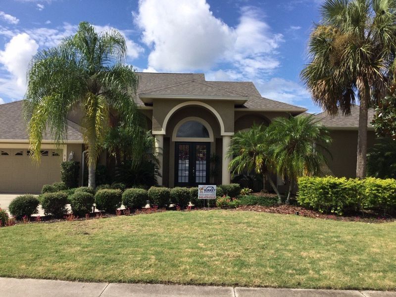 Tan house with arched doorway, palm trees, and green bushes against blue sky.