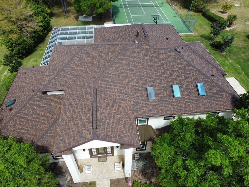 Aerial view of a brown shingle roof on a large house, with a tennis court and trees in the background.