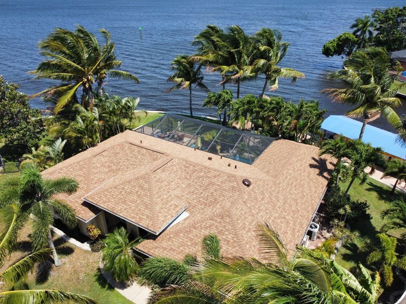 House with brown roof, pool, and palm trees by a body of water.