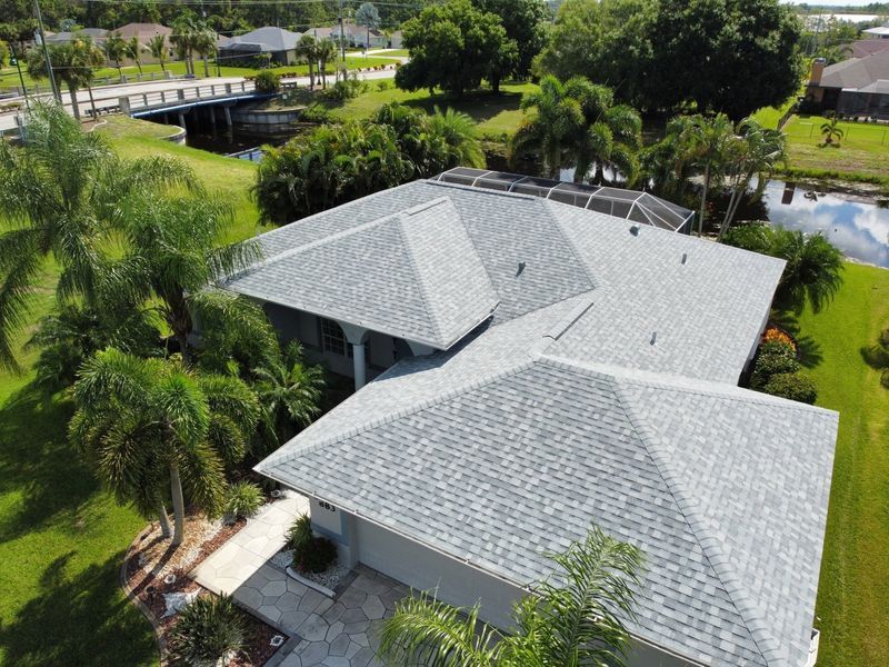 Aerial view of a gray asphalt shingle roof on a house surrounded by green trees and grass near a canal.