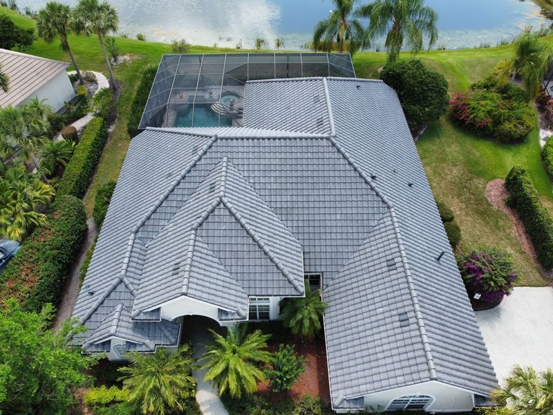 Aerial view of a gray tiled roof house with a screened pool, surrounded by green landscaping and palm trees.