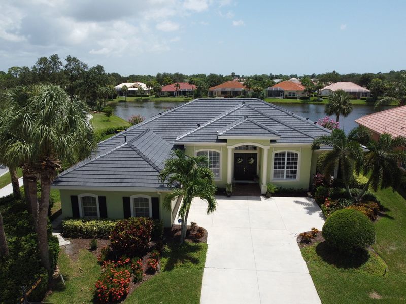 Aerial view of a light green house with a dark gray roof, palm trees, and a lake in the background.
