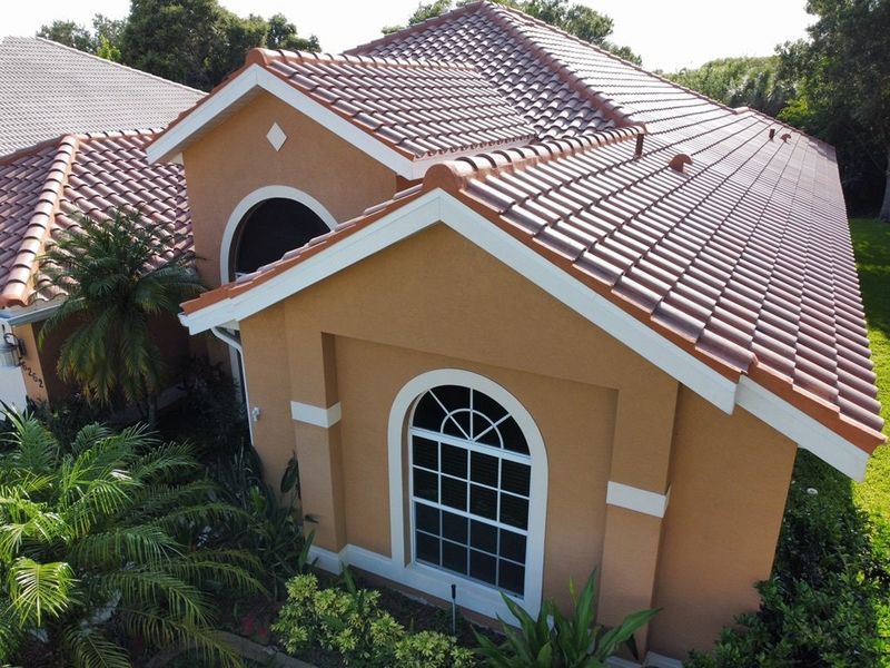 Tan house with clay tile roof and white trim, surrounded by green foliage.