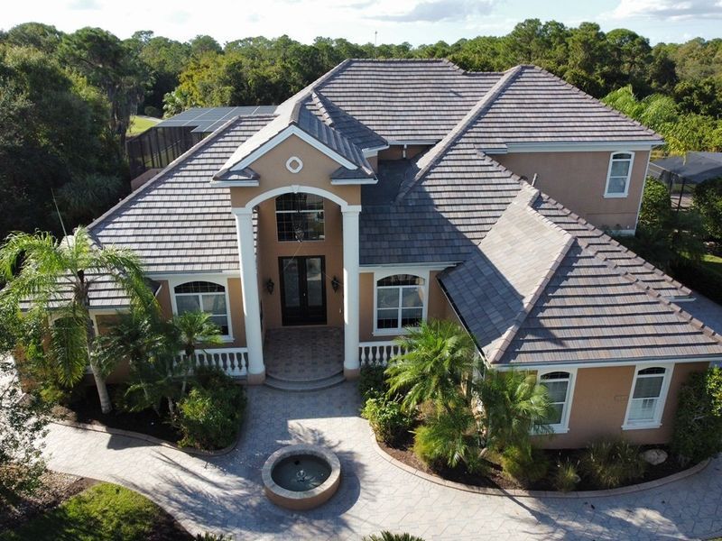 Large beige house with gray tile roof, circular driveway, fountain, and palm trees.
