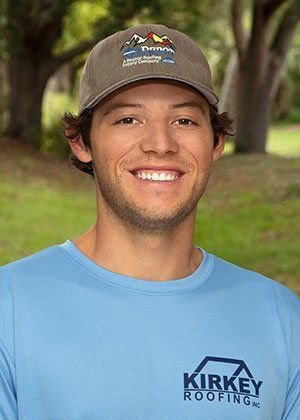 Man with a hat smiling, wearing a blue shirt with a roofing logo, standing outdoors.