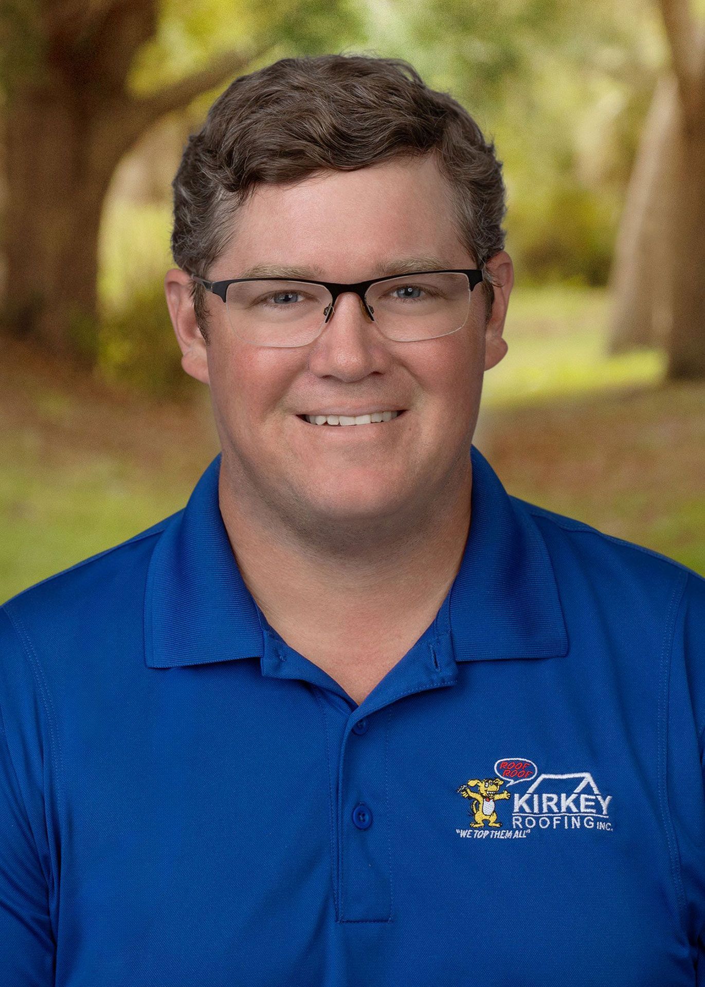 Man in blue polo shirt with glasses smiling; outdoors.