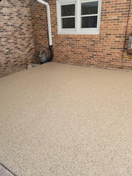 Tan speckled concrete patio against a brick wall with a window and white drainpipe.
