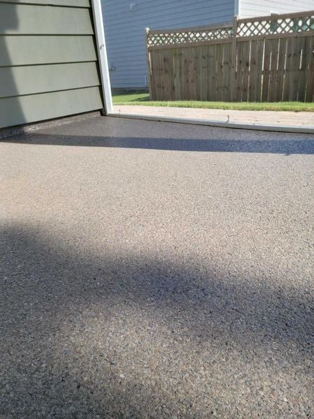 Asphalt driveway in sunlight, adjacent to a gray house exterior and a wooden fence.