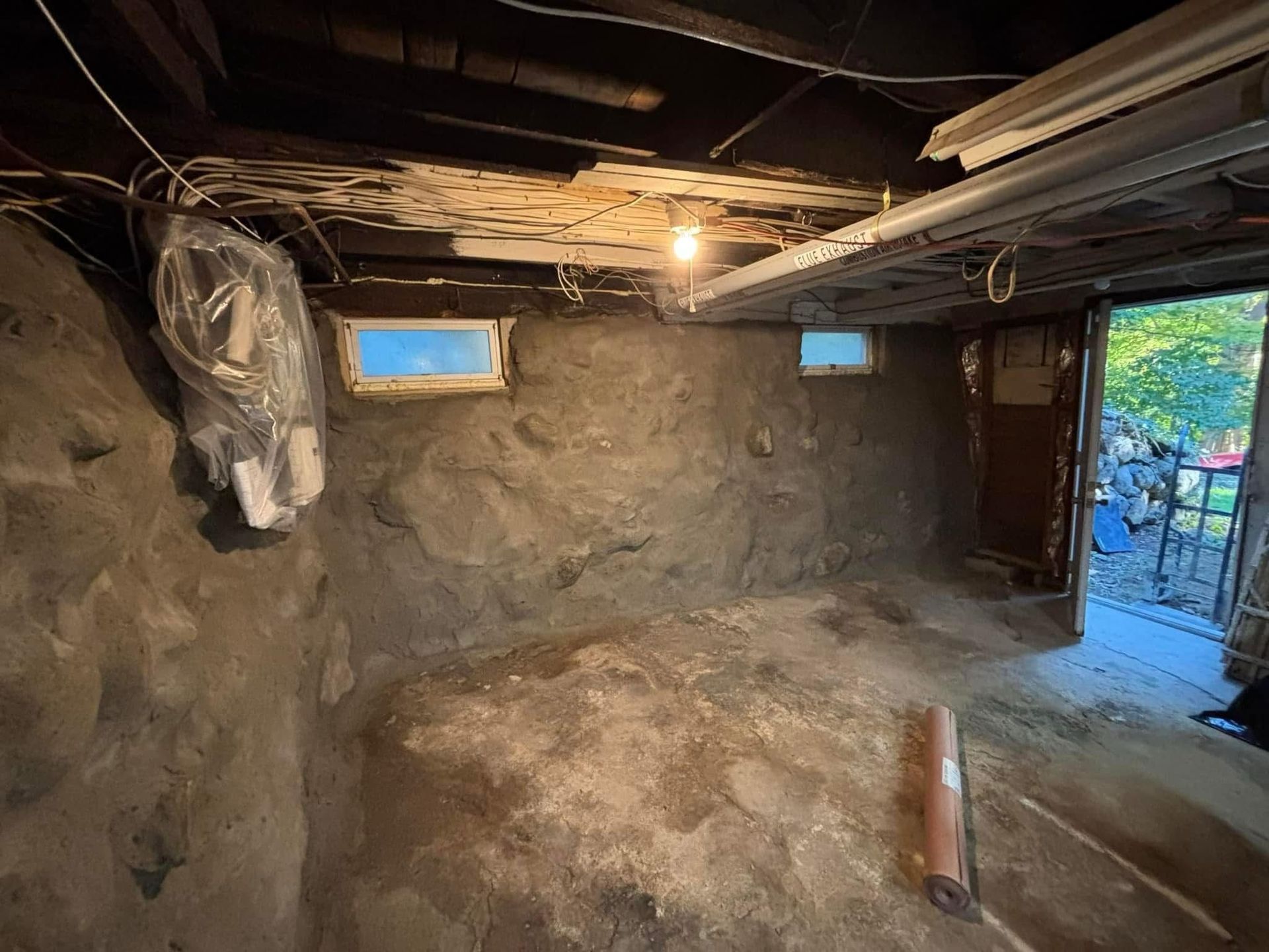 Small unfinished basement with dirt walls, exposed beams, two windows, and a single hanging light.