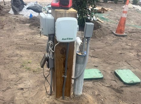 Electrical and irrigation control equipment mounted on a wooden post in a sandy, outdoor area.