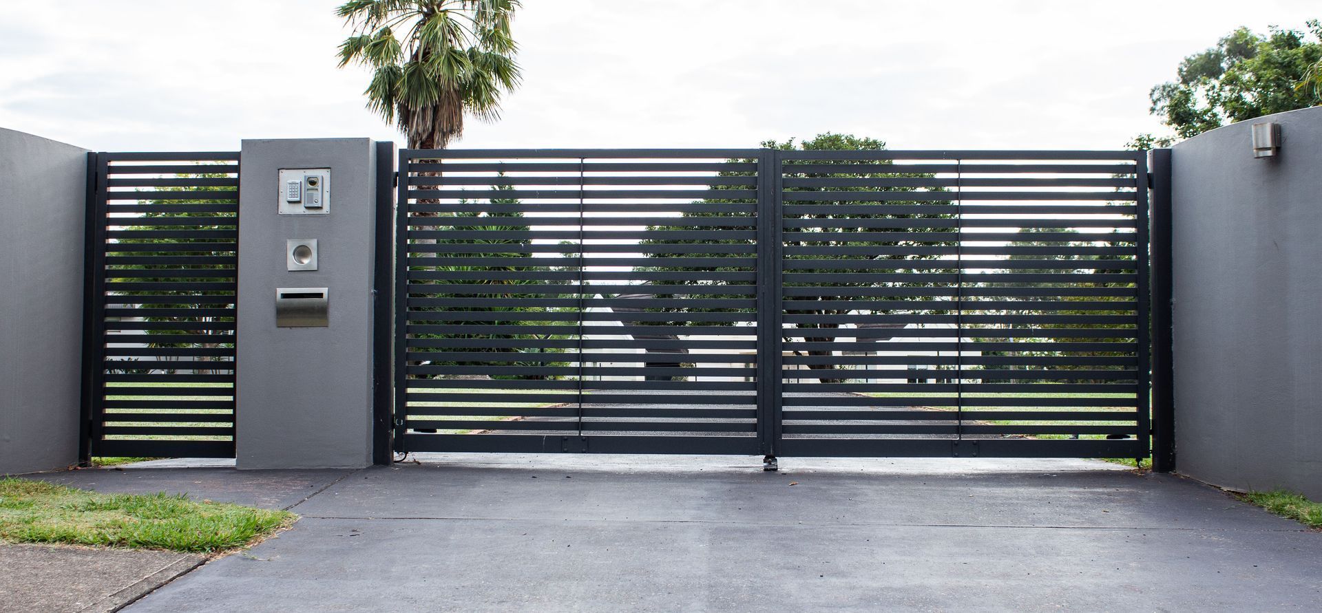 A gray driveway with a closed, black horizontal-slat metal security gate set between two gray concrete pillars.