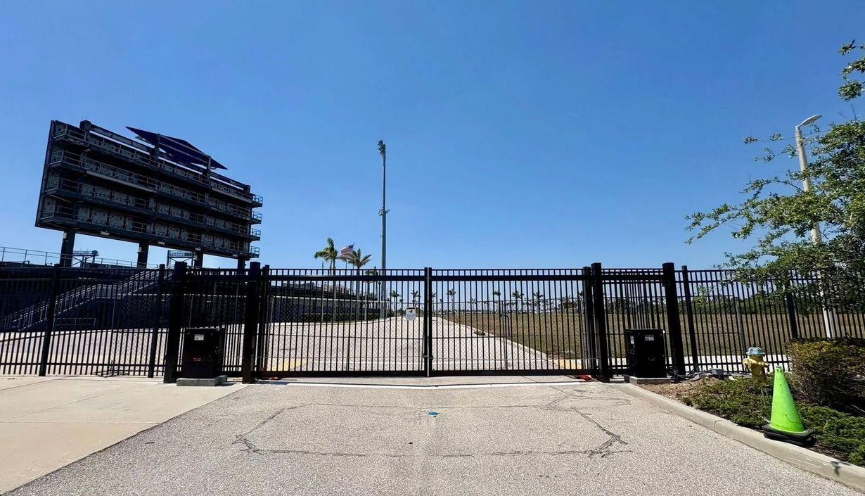 A black metal gate stands closed in front of a gravel lot, with a large, dark stadium scoreboard visible in the distance.