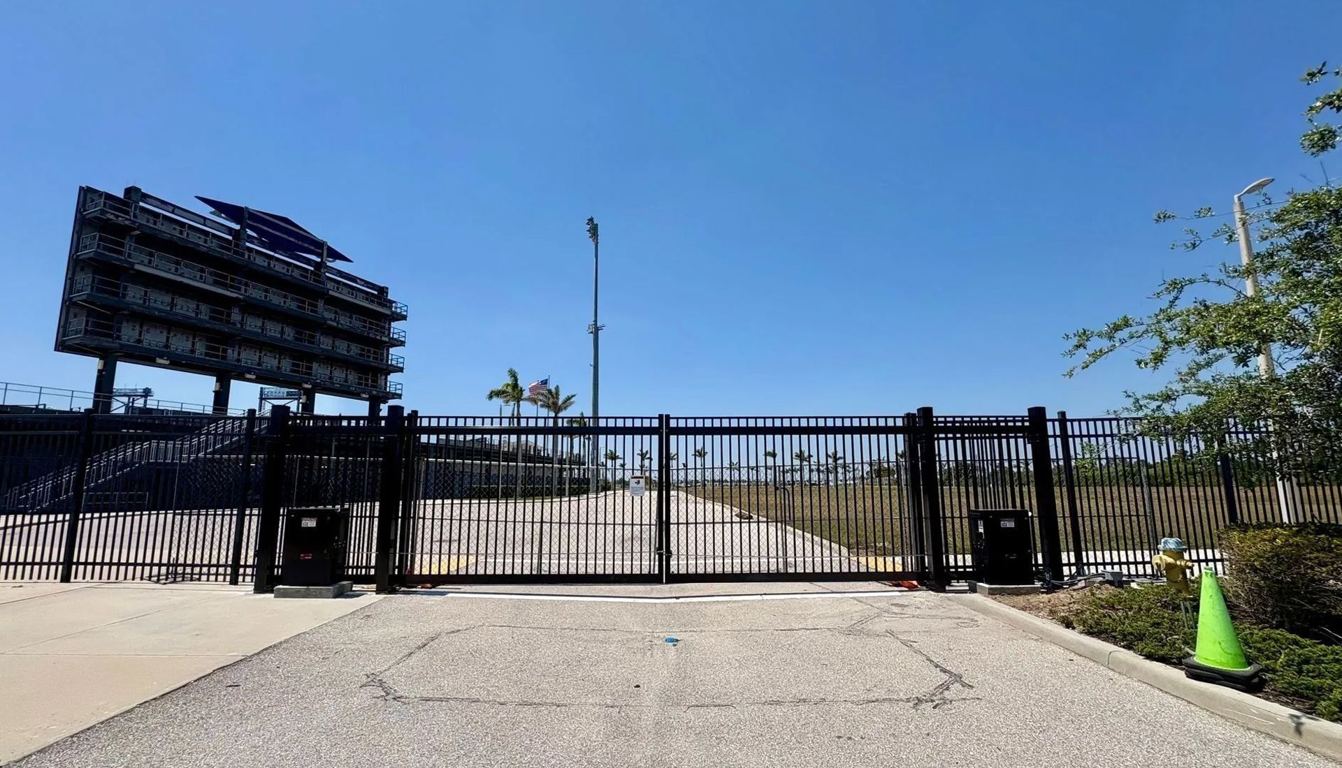 A black metal gate stands closed in front of a gravel lot, with a large, dark stadium scoreboard visible in the distance.