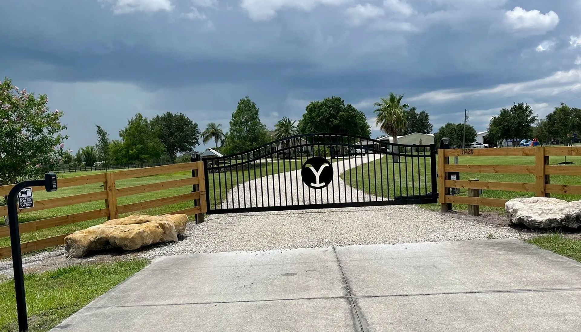 A driveway leads to a black metal gate featuring the Yellowstone ranch logo, set between wood fences under a cloudy sky.